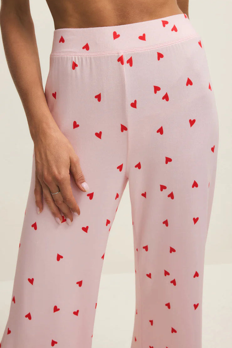 Pink pajama pants with red heart patterns worn by a person on a beige background