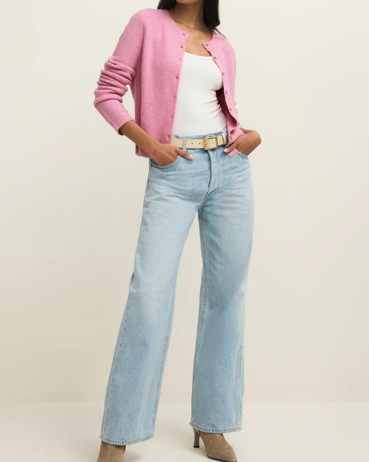 Woman wearing a pink cardigan, white top, light blue jeans, and beige shoes on a plain background