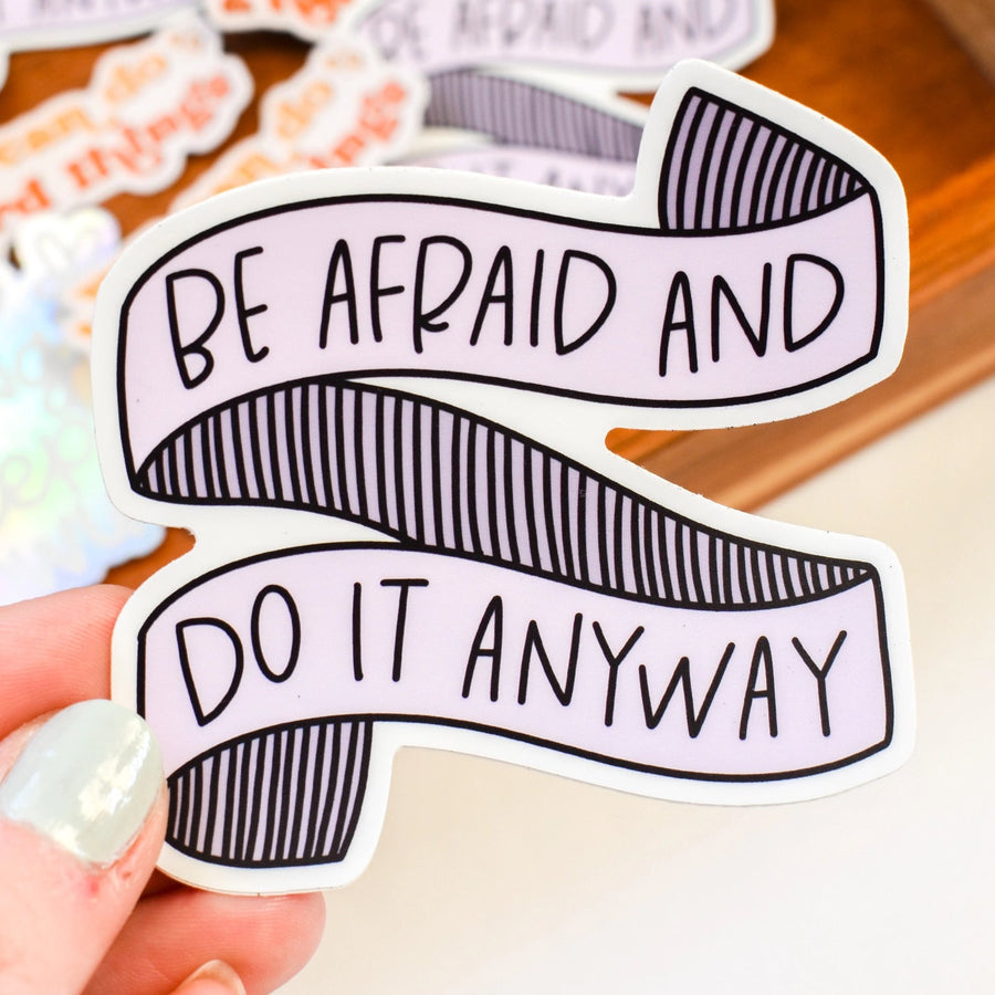Sticker with 'Be Afraid and Do It Anyway' text held by a hand on a blurred background